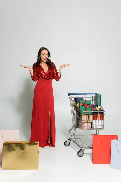 Opgewonden Vrouw Rode Jurk Zoek Naar Winkelwagen Met Cadeautjes Boodschappentassen — Stockfoto