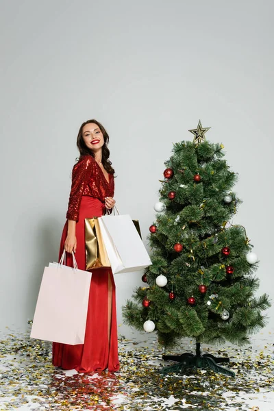 时尚女人一边拿着购物袋 一边在圣诞树旁和灰色背景的彩蛋旁对着相机微笑 — 图库照片
