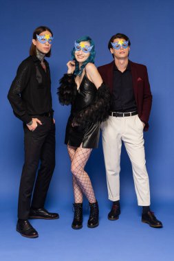 Mavi arka planda parti maskeleri ve şenlik kıyafetleriyle poz veren ikili olmayan insanların tam uzunluğu