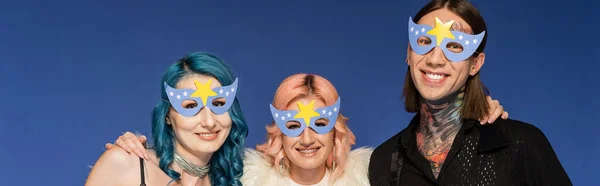 happy queer people in party masks smiling at camera isolated on blue, banner