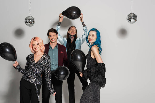 excited queer friends in festive clothes posing with black balloons on grey background