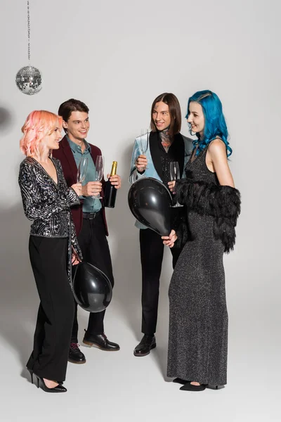 cheerful and elegant queer people with champagne glasses and black balloons on grey background