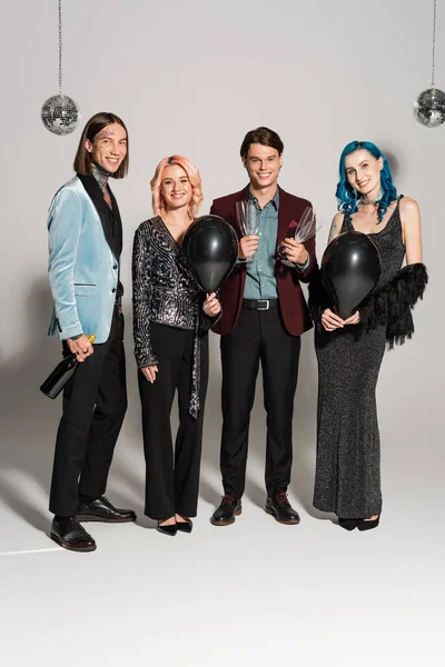 full length of happy queer people in festive clothes holding champagne and black balloons on grey background