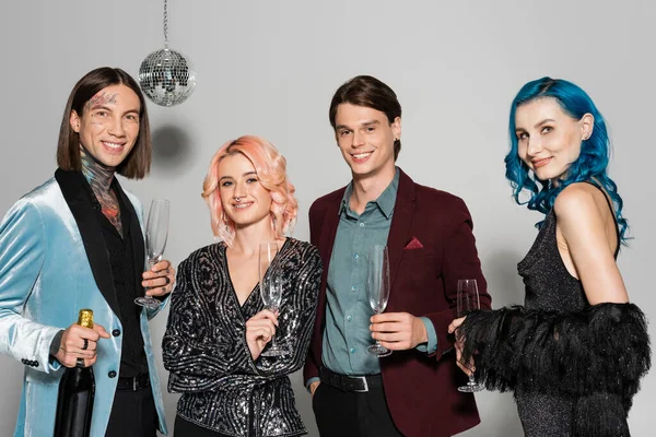 queer friends in festive clothes holding champagne glasses and looking at camera on grey background