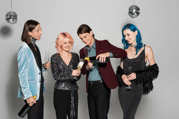 улыбающийся педик разливает шампанское во время рождественской вечеринки с элегантными небинарными друзьями на сером фоне
