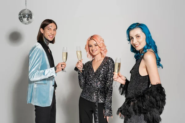 elegant queer people with glasses of champagne smiling at camera while celebrating christmas on grey background