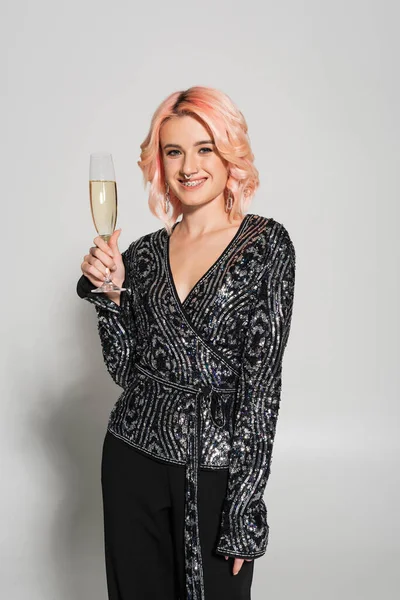 elegant woman in dental braces smiling at camera and holding champagne glass on grey background