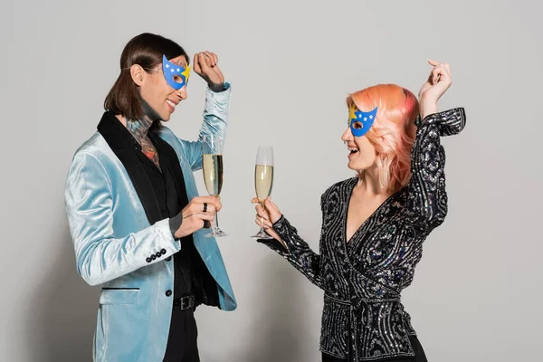 stylish and happy queer people in party masks dancing with champagne glasses on grey background