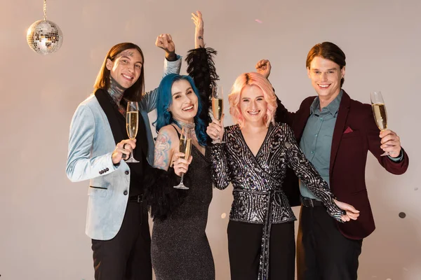 cheerful nonbinary people with champagne glasses and raised hands on beige background