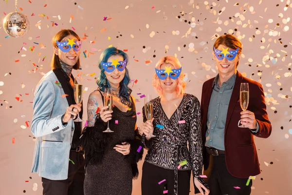 happy and elegant queer friends in party masks holding champagne glasses under falling confetti on beige background
