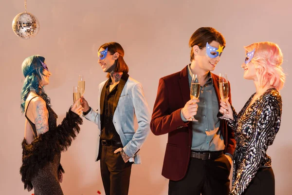 elegant nonbinary people in party masks holding champagne glasses and talking on beige background