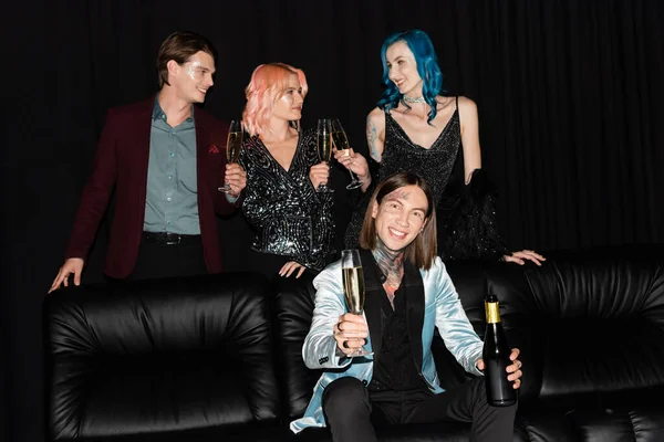 tattooed queer man sitting on couch with bottle of champagne near nonbinary friends clinking glasses on black background