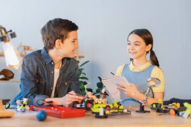 Gülümseyen kız evde arkadaşının ve robot modelinin yanında defter tutuyor. 