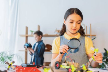 Evdeki masada elinde büyüteç ve robot modeli tutan çocuk. 