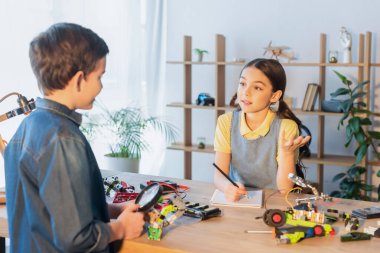 Not defteri ve kalemi olan genç bir kız robot modelinin parçaları yanında elinde büyüteçle arkadaşıyla konuşuyor.