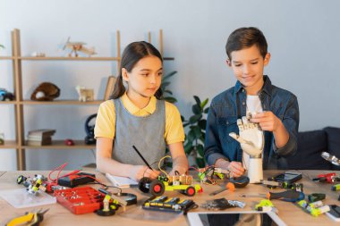Kız deftere yazarken oğlan mekanik parçaların yanında robot el tutuyor ve dijital tableti bulanıklaştırıyor.