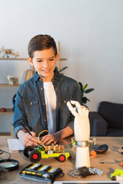 Mutlu çocuk robotlara bakıyor. Aletlerin yanında ve evdeki masada mekanik parçalar var.