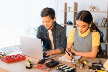 Mutlu çocuk dizüstü bilgisayar kullanırken kız robotik modelin detaylarını not alıyor.