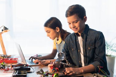 Gülümseyen çocuk elinde büyüteçle kızın yanında dizüstü bilgisayar kullanarak robot modeli birleştiriyor.
