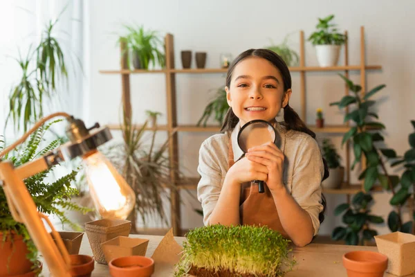 Smiling kid holding magnifying glass near blurred plants and lamp at home