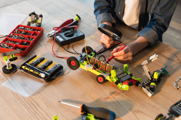 Cropped view of boy holding magnifying glass near robotic model and millimeter on table at home 