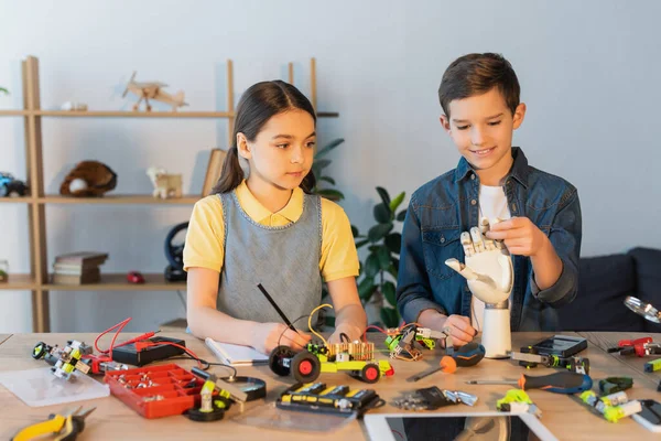 Flicka Skriva Anteckningsbok Medan Pojke Håller Robotteknik Hand Nära Mekaniska — Stockfoto