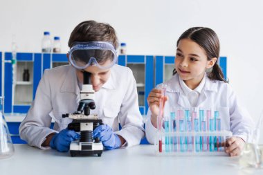 Arkadaşının yanında test tüpü tutan gülümseyen kız kimyasal deney sırasında mikroskoba bakıyor. 