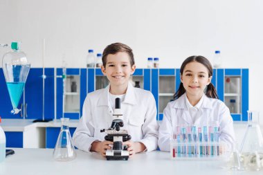 Beyaz önlüklü mutlu çocuklar mikroskop yakınındaki kameraya ve mataralı test tüplerine bakıyor.