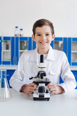 Laboratuvarda mikroskop yakınında kameraya bakan beyaz önlüklü mutlu çocuk.