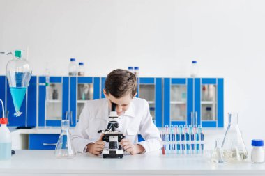 Beyaz önlüklü çocuk laboratuarda test tüpleri ve mataraların yanında mikroskoba bakıyor.