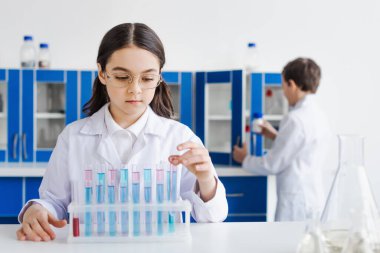 Beyaz önlüklü bir kız ve test tüplerinin yanında gözlüklü sıvı ve arka planda erkek çocuk.
