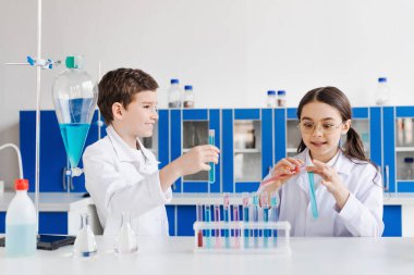 Beyaz önlüklü mutlu çocuklar mikroskop yakınındaki kameraya ve mataralı test tüplerine bakıyor.