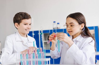 Gözlüklü kız test tüplerinde sıvıyı karıştırıyor. Yanında beyaz önlüklü gülen bir arkadaş var.