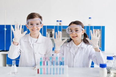 Beyaz önlüklü ve gözlüklü gülümseyen çocuklar lateks eldivenli ellerini laboratuvar tüplerinin yanında gösteriyor.