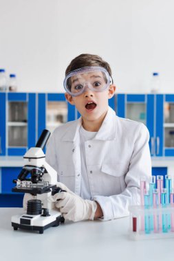 Gözlüklü çocuğun mikroskobun yanındaki kameraya ve laboratuardaki test tüplerine bakması hayret verici.
