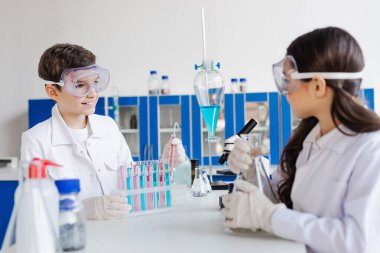 Gözlüklü çocuk test tüplerinin yanında bulanık kıza ve laboratuardaki şişelere bakıyor.