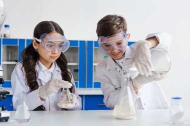 tensed girl in goggles looking at smiling boy pouring liquid into flask while doing chemical experiment clipart