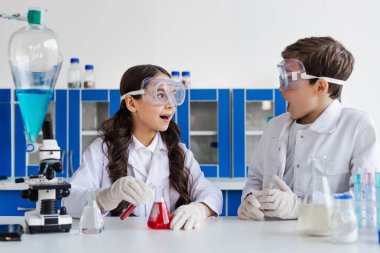 Gözlüklü kız kimya laboratuvarında kırmızı sıvıyla mataranın yanındaki arkadaşına bakıyor.