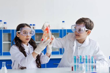 Beyaz önlüklü mutlu çocuklar mikroskop yakınındaki kameraya ve mataralı test tüplerine bakıyor.