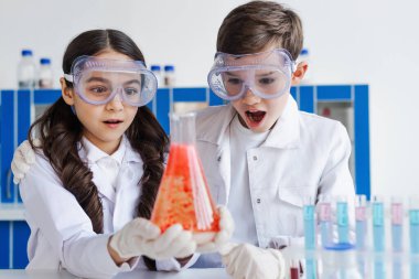Gözlüklü arkadaşlar laboratuarda kırmızı maddeyle mataraya bakıyorlar.