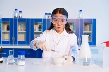 Kirli suratlı, kasenin yanında cımbız ve laboratuarda buharlı matara tutan sürpriz kız.