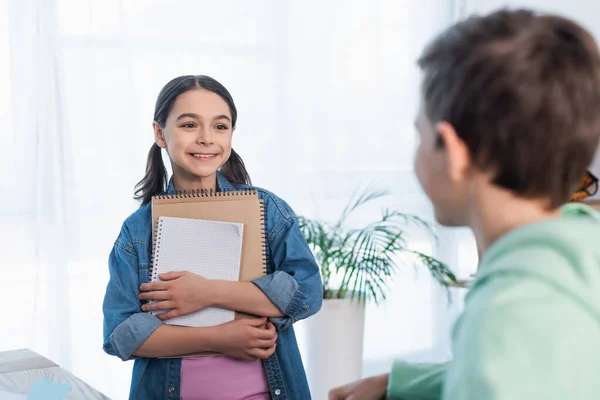 Cheerful Girl Holding Notebooks Looking Blurred Boy Home — Stock Photo, Image
