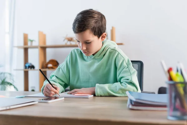 专心致志的男孩在接近模糊的手机的抄袭本上做作业和写作 — 图库照片