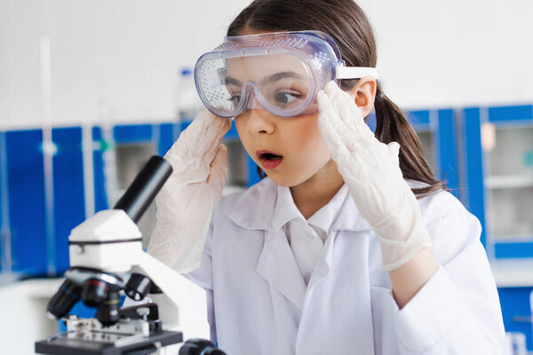 shocked girl in goggles and latex gloves touching head near microscope in laboratory