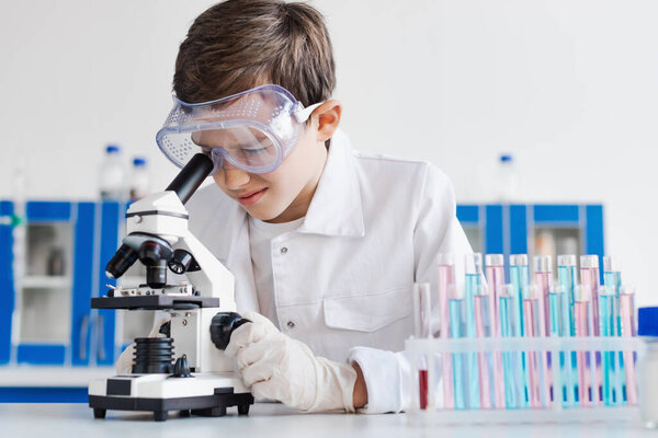 preteen kid in goggles and latex gloves looking into microscope near test tubes 
