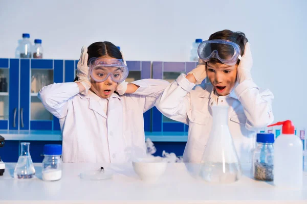 astonished children with dirty faces touching heads while looking at steaming bowl in chemical lab