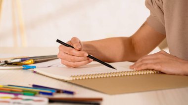 Bulanık renkli kalemlerin yanındaki çizim defterine çizim yapan kadının kırpılmış görünümü 