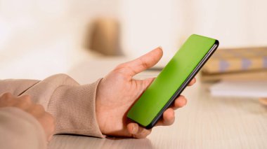 Evde elinde yeşil ekranlı cep telefonu olan bir kadının kısmi görüntüsü 