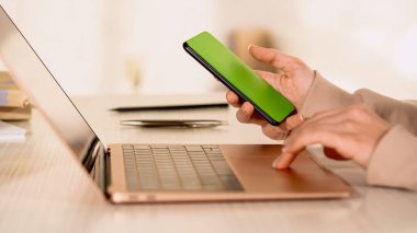 Akıllı telefonu boş ekranla tutan ve evde dizüstü bilgisayar kullanan bir kadın görüntüsü. 