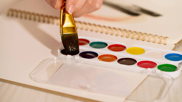 Cropped view of woman holding paintbrush near watercolor paint and sketchbook 